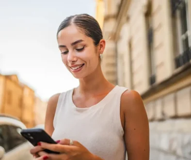 Mladá žena se usmívá na ulici do telefonu.