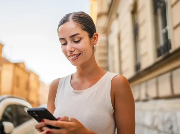 Mladá žena drží telefon v ruce a usmívá se.