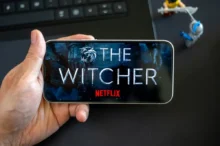 Ruka s telefonem, kde je logo Netflixu a The Witcher. Pod ním jsou postavičky.