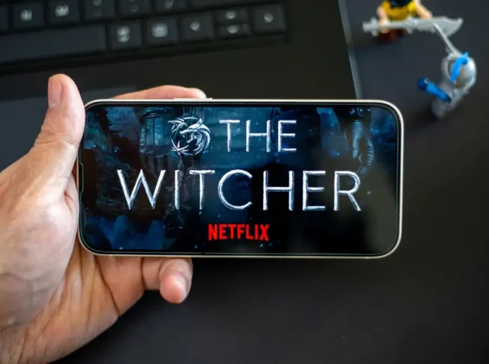 Ruka s telefonem, kde je logo Netflixu a The Witcher. Pod ním jsou postavičky.