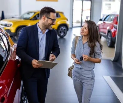 Muž v saku mluví s ženou uprostřed automobilové prodejny.