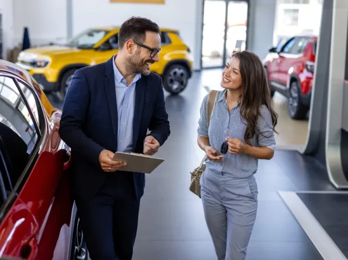 Muž v saku mluví s ženou uprostřed automobilové prodejny.